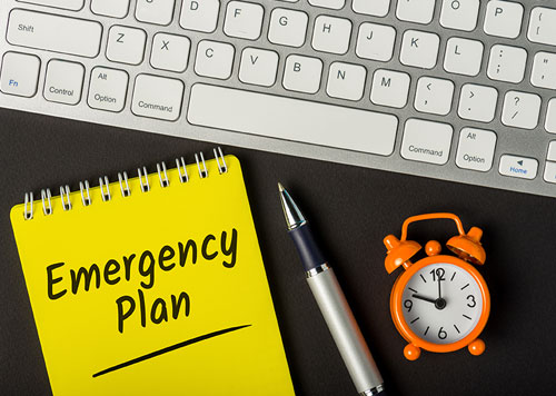 Emergency Plan writing on a yellow notepad placed next to a pen, alarm clock & keyboard