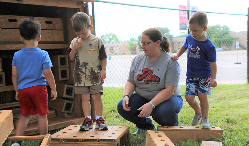 Three little boys and a teacher play with large hallow wooden outdoor blocks.